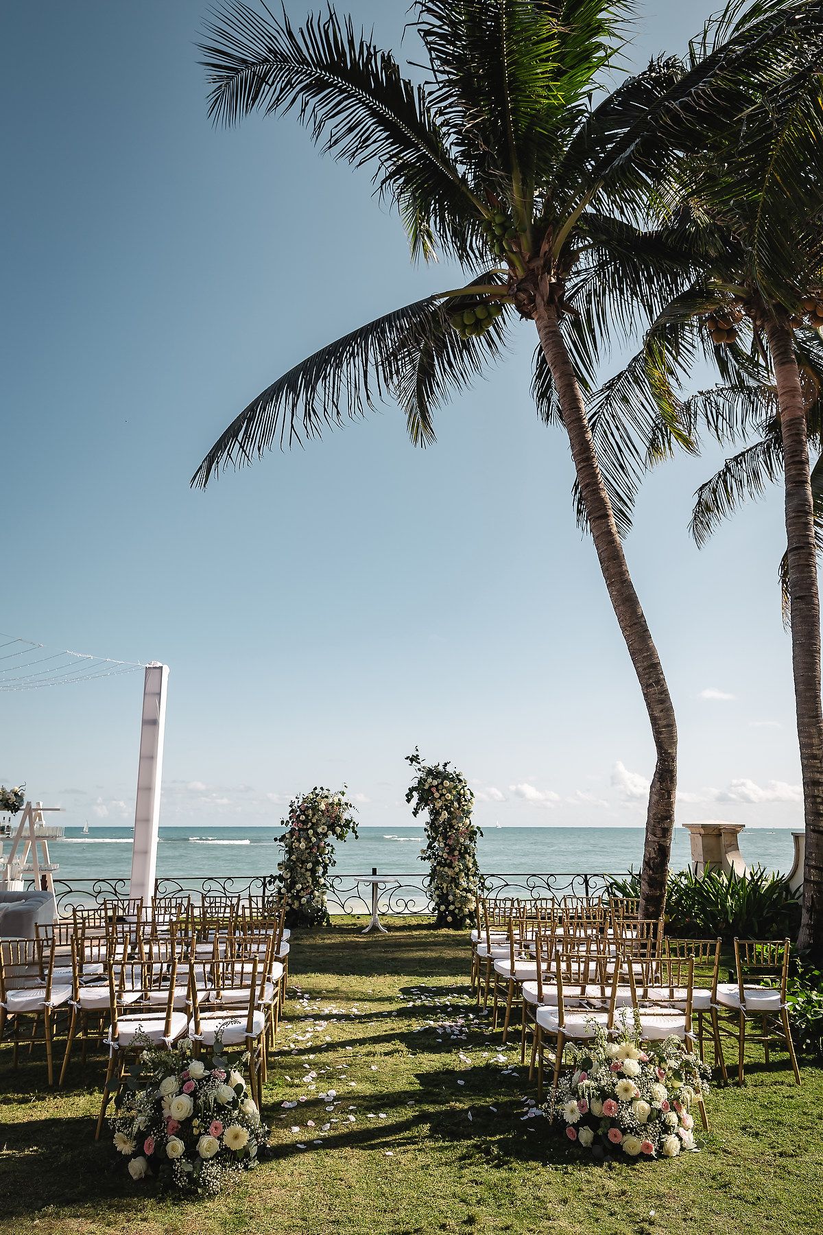 Beachfront wedding ceremony setup with white chairs and palm trees overlooking the ocean.