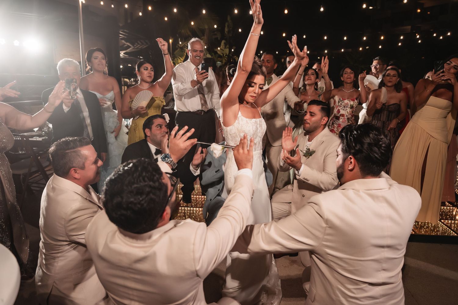 Energetic wedding party dancing and celebrating under warm string lights at an evening reception.