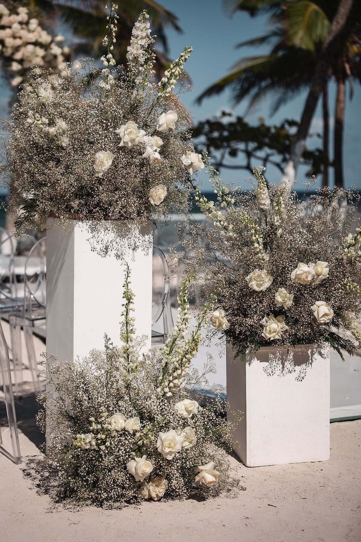 White floral arrangements and decorative elements line a beach wedding aisle with palm trees in the background.