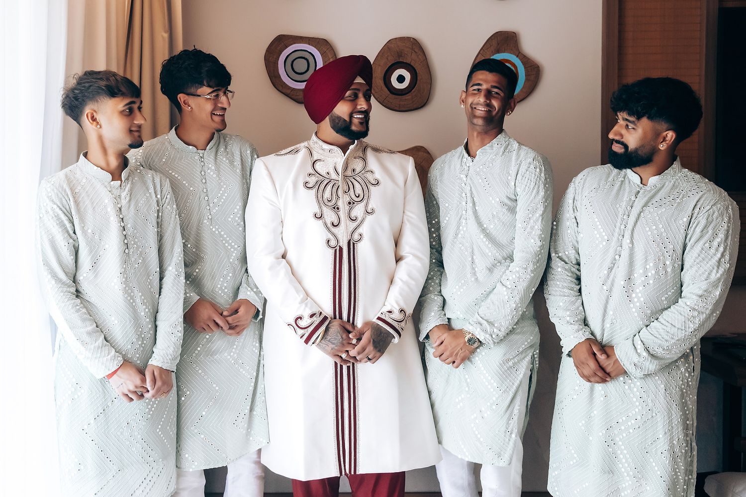 Traditional Indian wedding party wearing elegant white kurtas and burgundy sherwani standing against a wall.