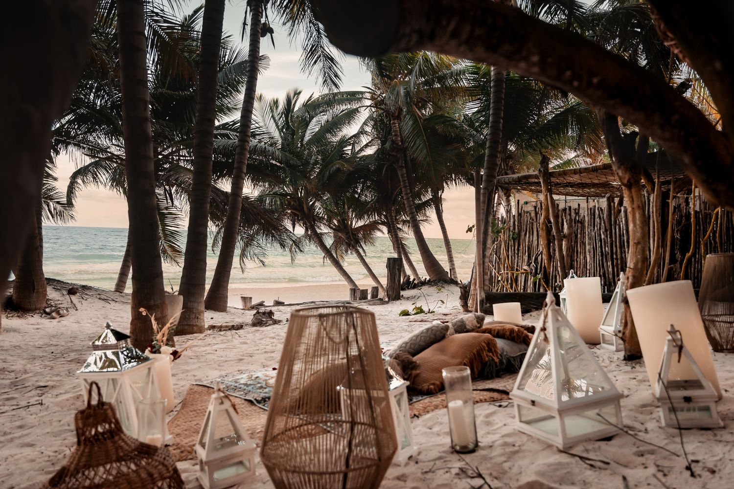 Romantic beach setup with wicker chairs and lanterns among palm trees at sunset.