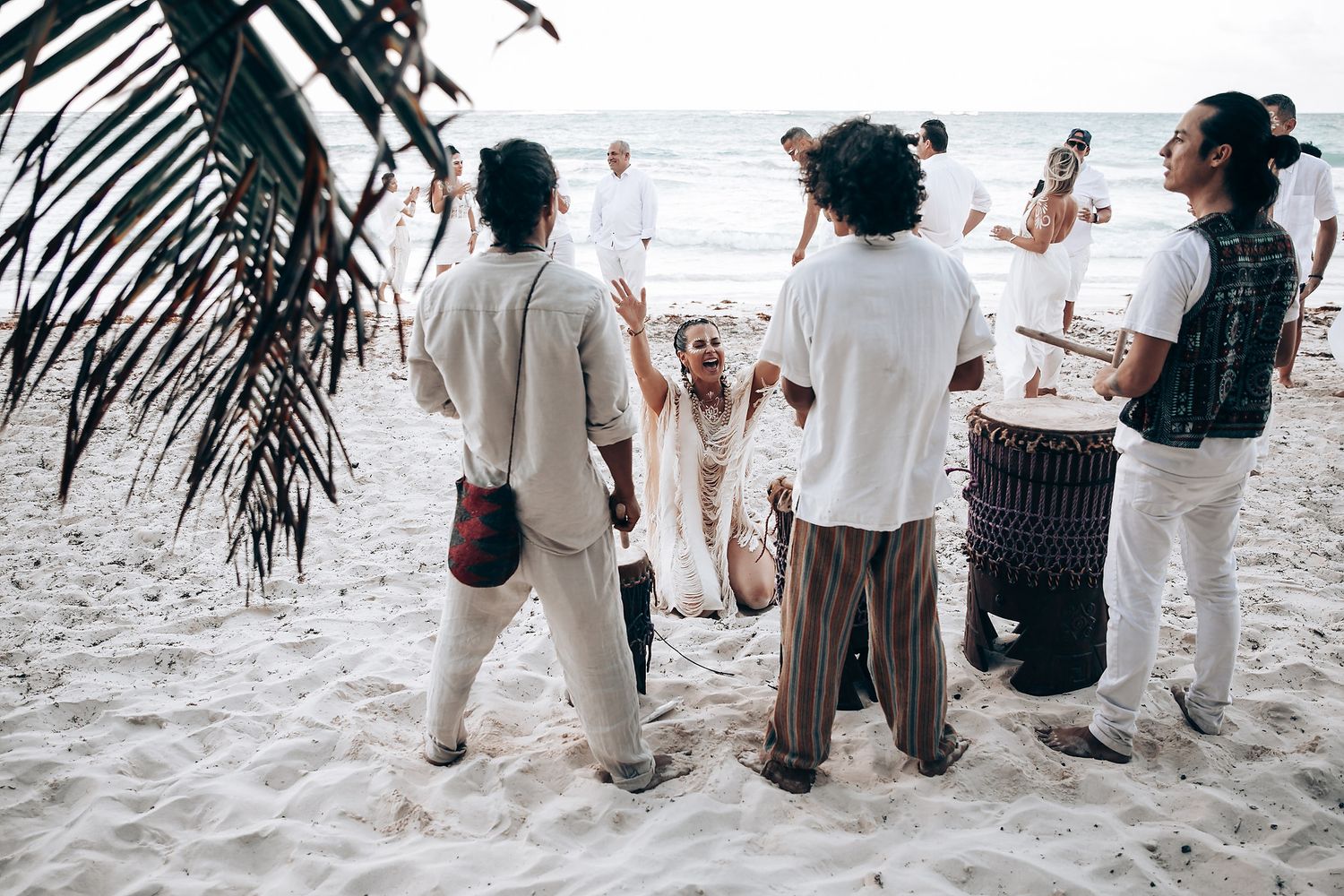 Wedding attendees in white clothing stand together on a tropical beach with palm trees.