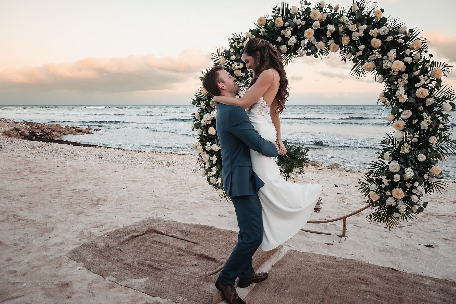 Groom lifting bride in white dress by floral arch on beach at sunset during romantic outdoor wedding ceremony.