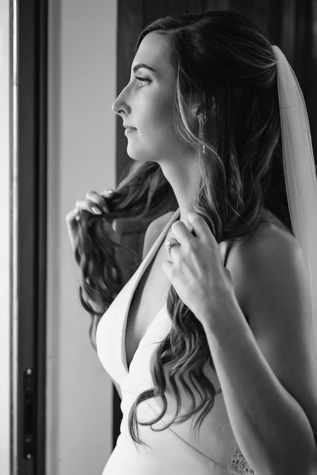Black and white portrait of a bride in a white gown and veil, adjusting her hair near a window.