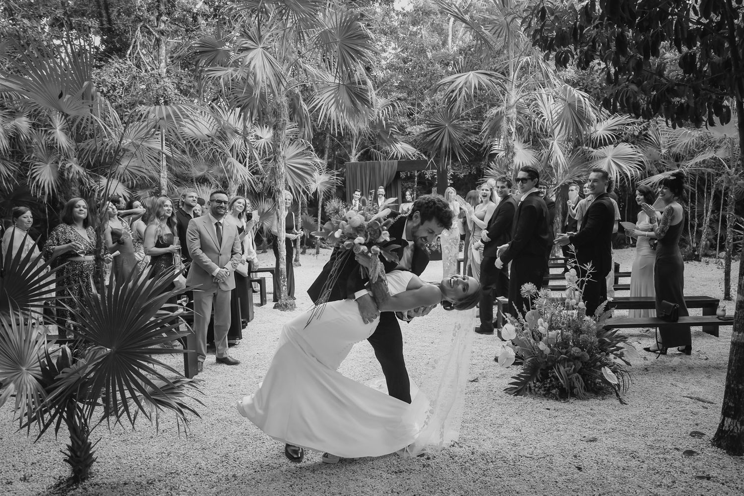 Bride and groom share a romantic dip kiss at outdoor tropical wedding ceremony in black and white.