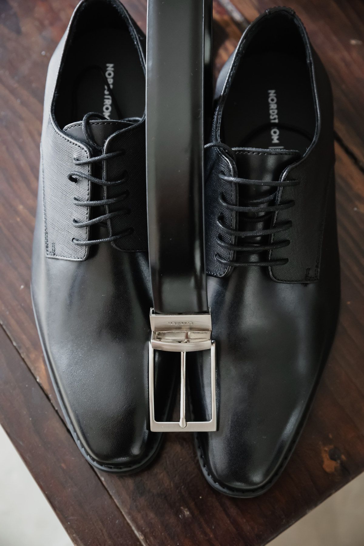 Black leather dress shoes with laces and a black belt with silver buckle displayed on a wooden surface.