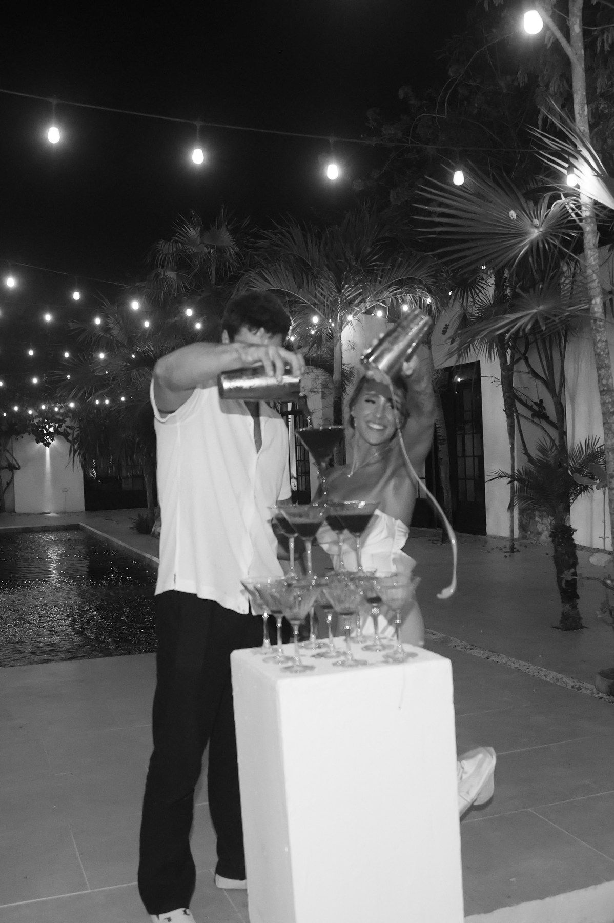 Couple pouring champagne tower outdoors at night with string lights and palm trees in black and white photo.