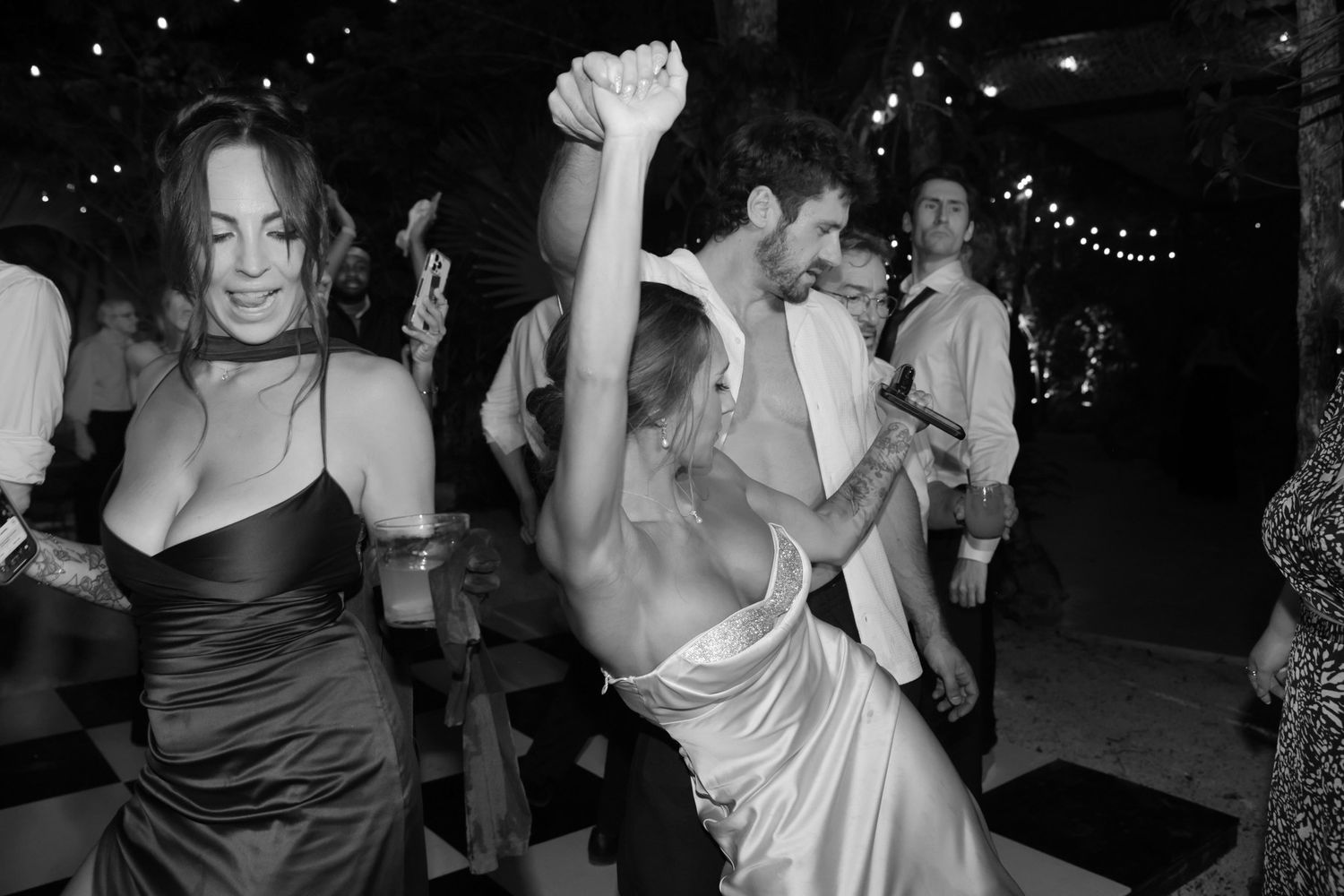 Black and white photo of guests dancing joyfully at a wedding reception under string lights.