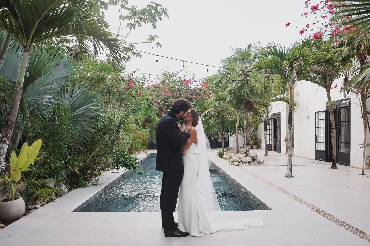 Bride and groom share a romantic kiss beside a reflecting pool surrounded by tropical palms.