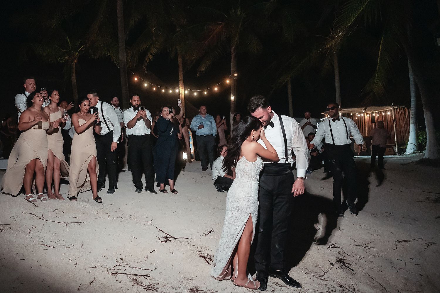 Bride and groom share first dance on sandy beach at night with string lights and guests applauding at outdoor wedding reception.