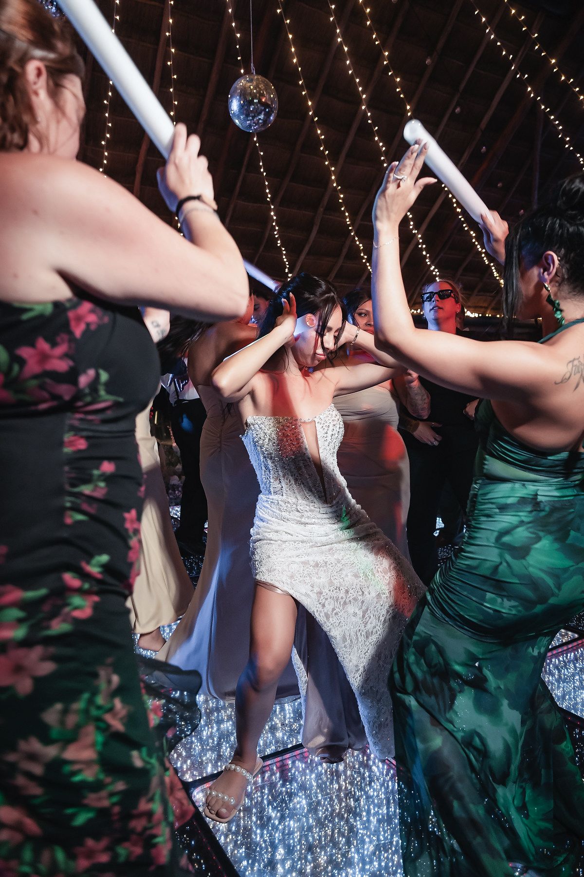 Bride in white gown dancing with guests at wedding reception under disco ball and string lights.