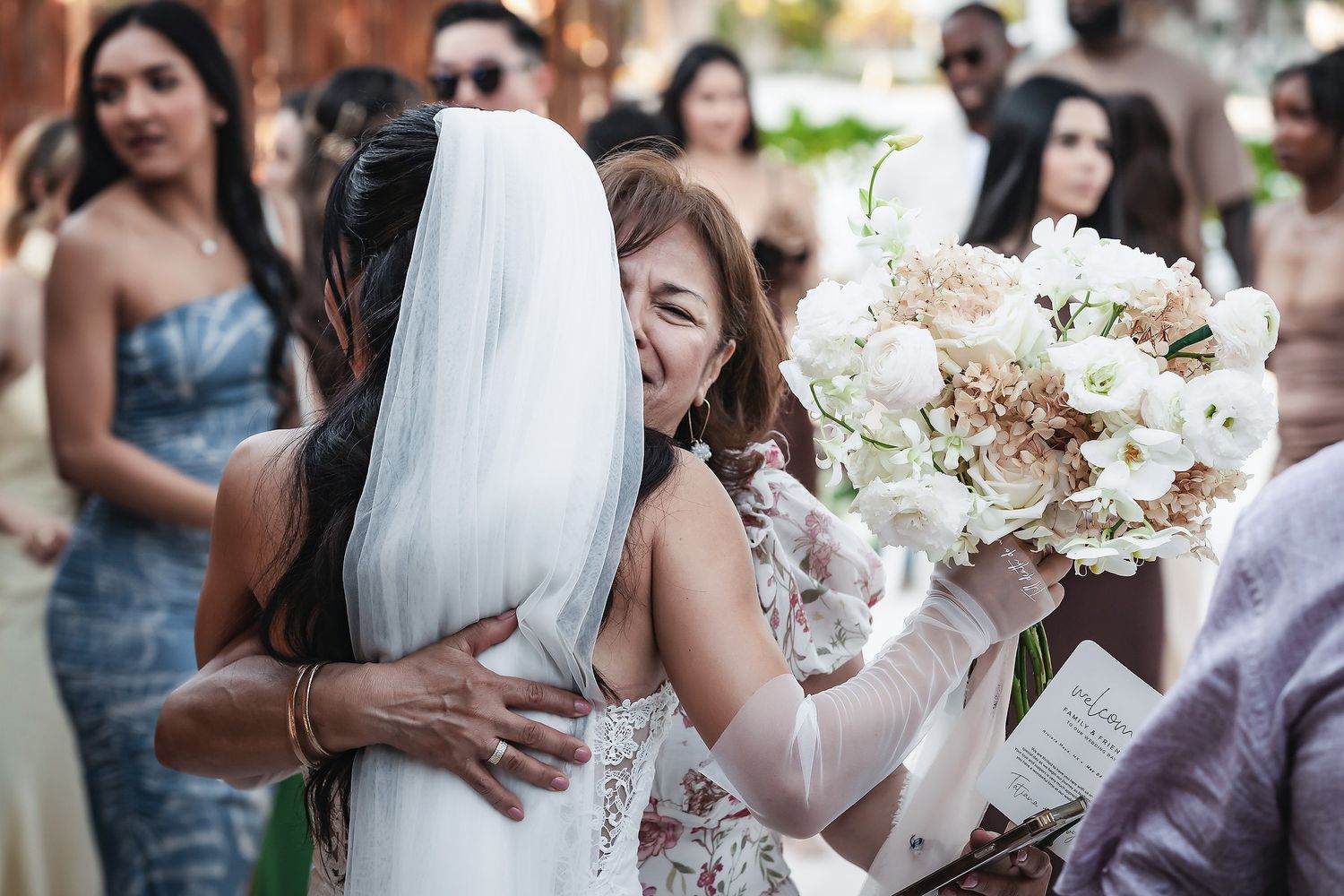 Bride in white veil hugging a smiling woman holding a bouquet of white and blush flowers at a wedding.