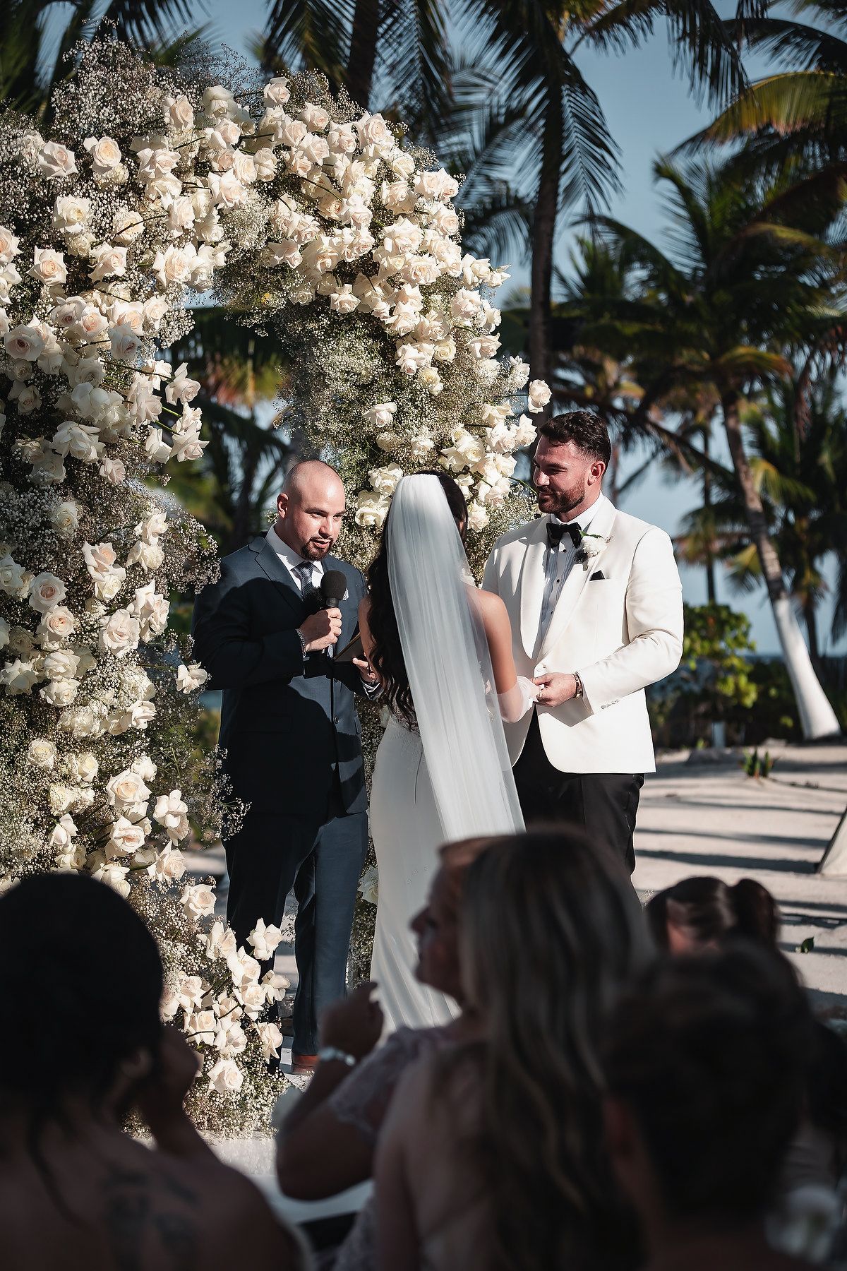 Outdoor beach wedding ceremony with groom in white tuxedo, bride in veil, officiant, and floral arch of white roses and palms.