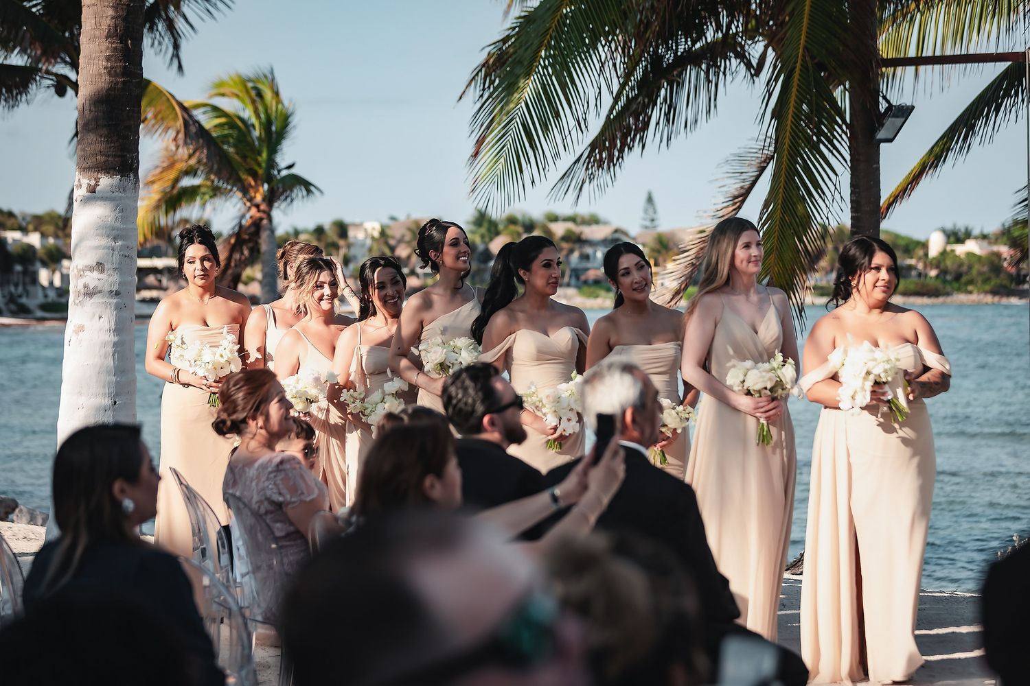 Bridesmaids in blush gowns holding white bouquets stand outdoors near palm trees and water at a tropical wedding ceremony.