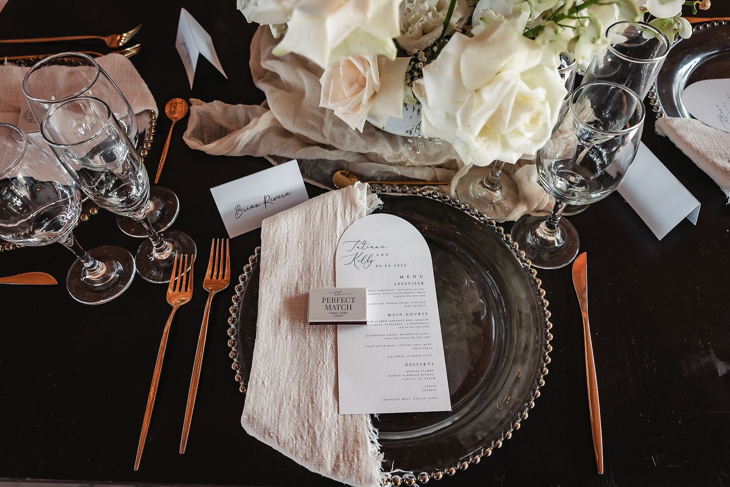 Elegant wedding table setting with gold cutlery, crystal glasses, white floral centerpiece, and a menu card on a beaded charger plate.