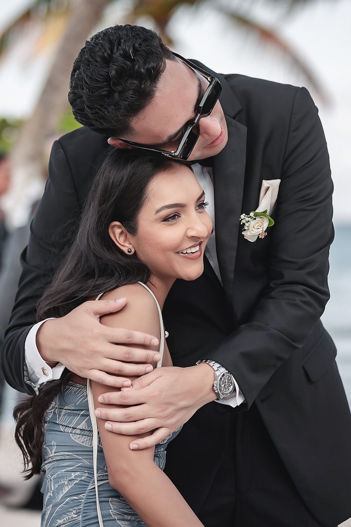 Groom in black suit embraces bride in blue dress during outdoor wedding celebration near palm trees.