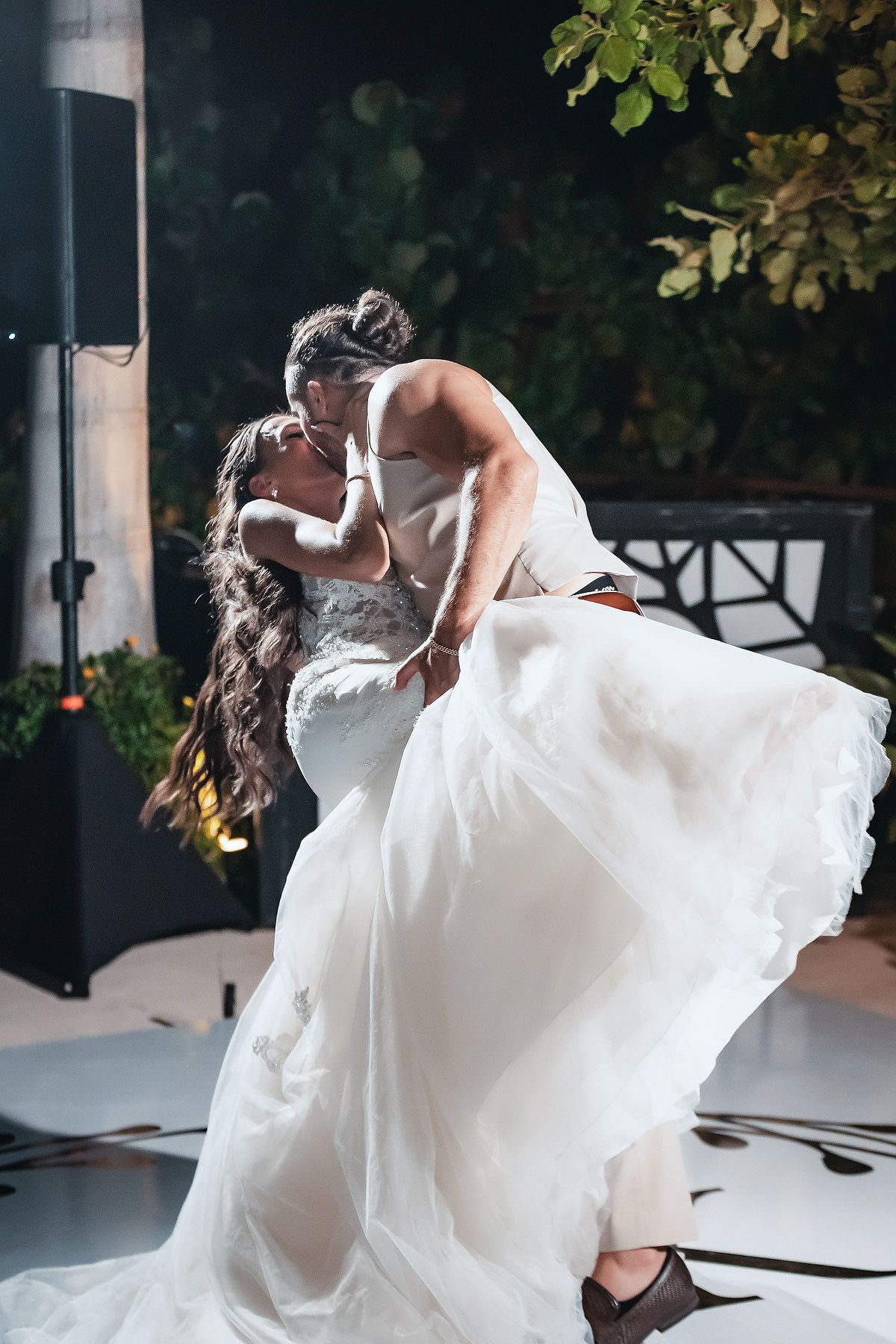 Groom lifts bride in wedding dress during passionate first dance kiss at outdoor reception.