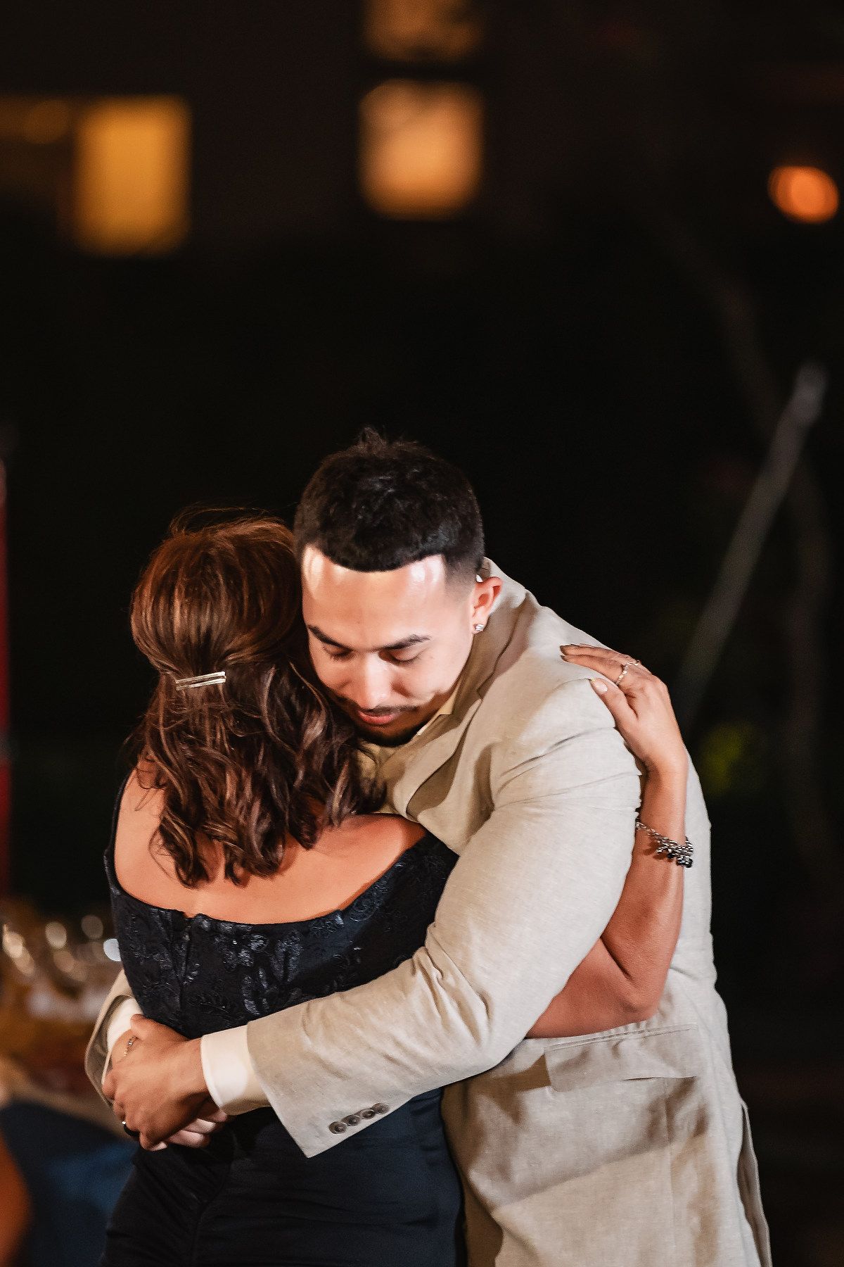 A groom in a beige suit shares an emotional dance with a woman in a black dress at a wedding reception.