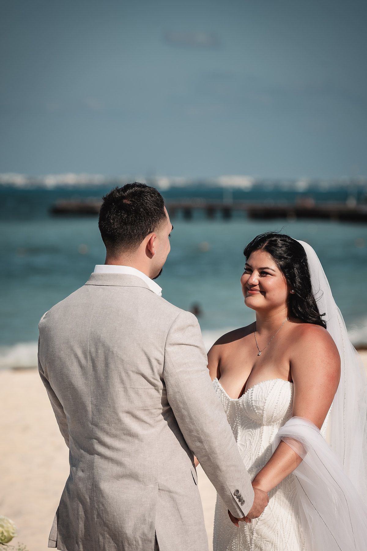 Bride and groom exchange vows on a beach, with turquoise ocean waters visible in the background.