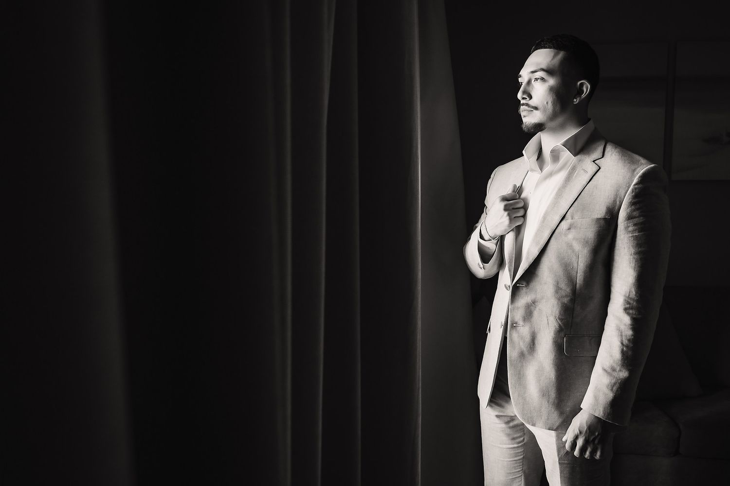 Black and white photo of a stylish man in a suit adjusting his tie near a curtained window.