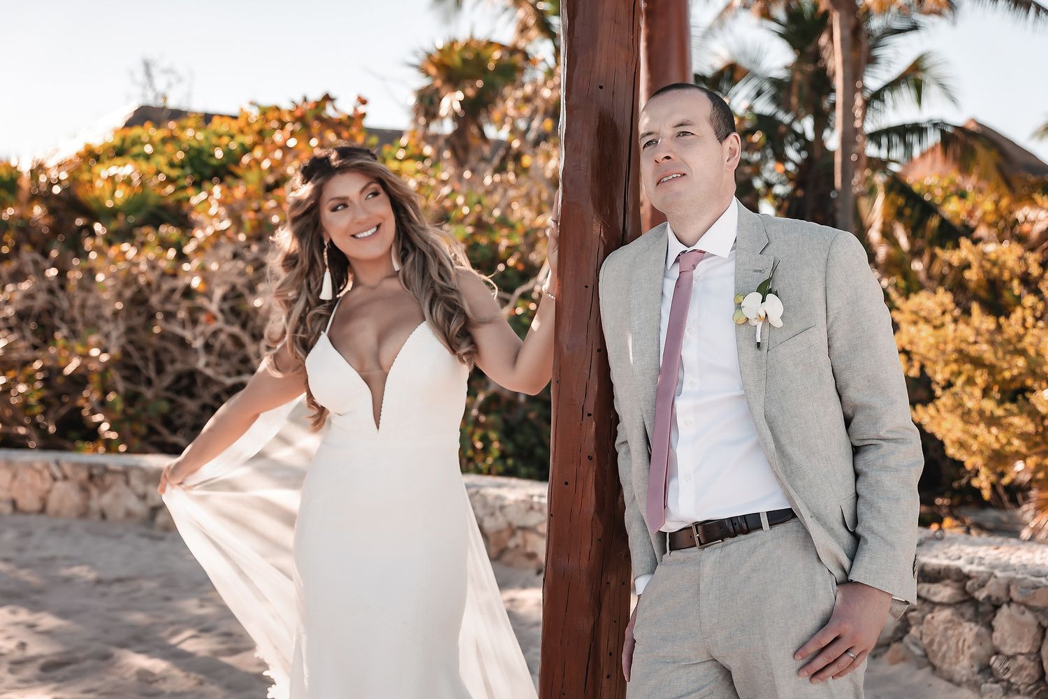 Bride in white gown and groom in tan suit with pink tie pose outdoors at a tropical beach wedding venue.