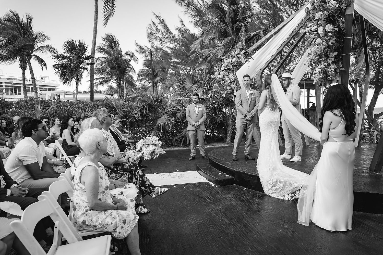 Black and white outdoor beach wedding ceremony with couple at altar, guests seated, palm trees and ocean in background.