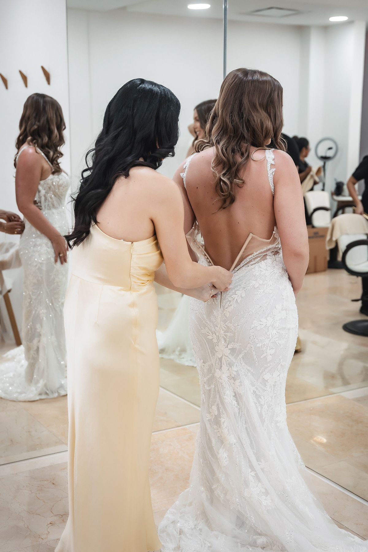A bridesmaid in yellow helps a bride in a lace backless gown in a bridal boutique fitting room.