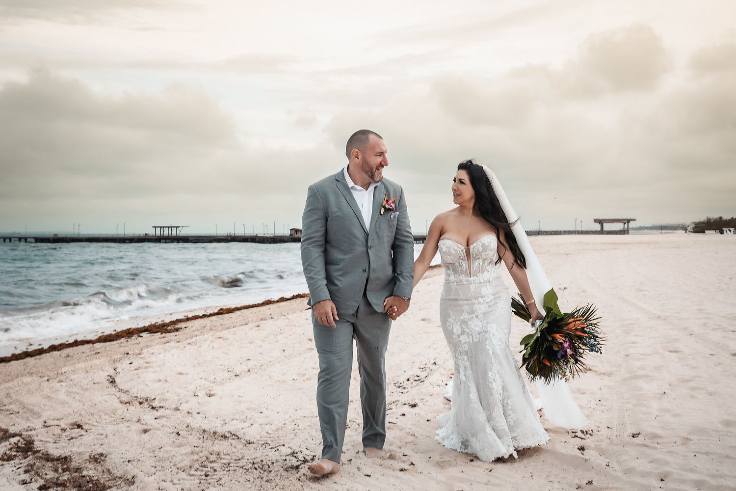 Bride and groom walking hand in hand on a sandy beach near a pier, bride holding a vibrant bouquet in a white gown.