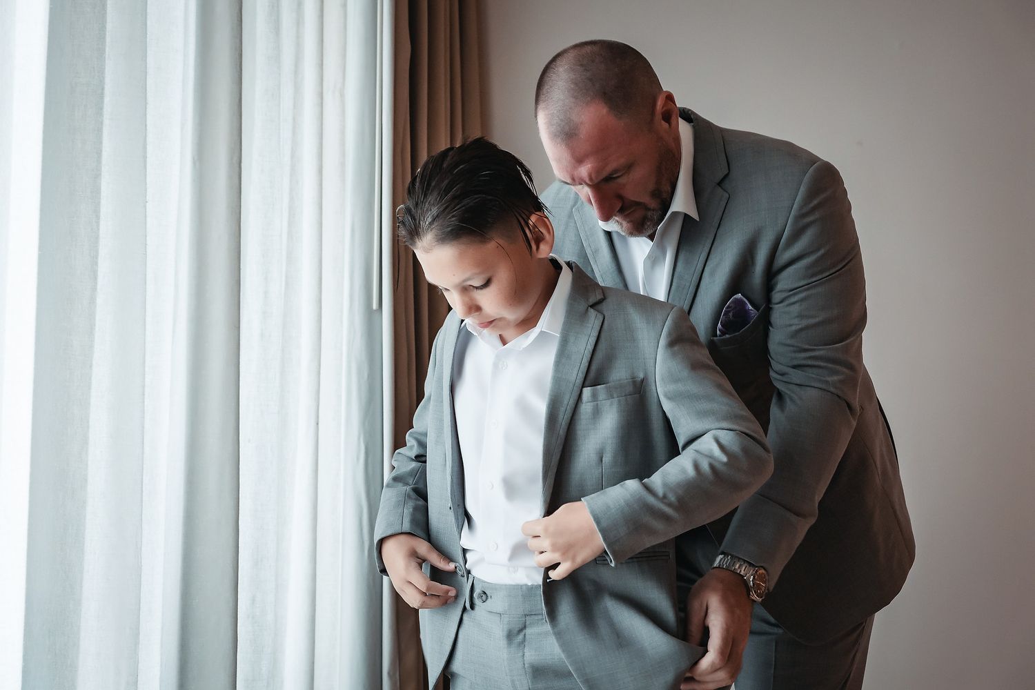 An older man in a gray suit helps a young boy adjust his matching gray suit jacket near a window.