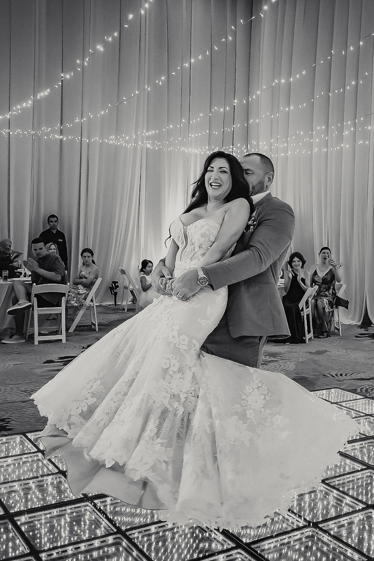 Bride and groom share their first dance on a mirrored floor beneath twinkling fairy lights at elegant wedding reception.
