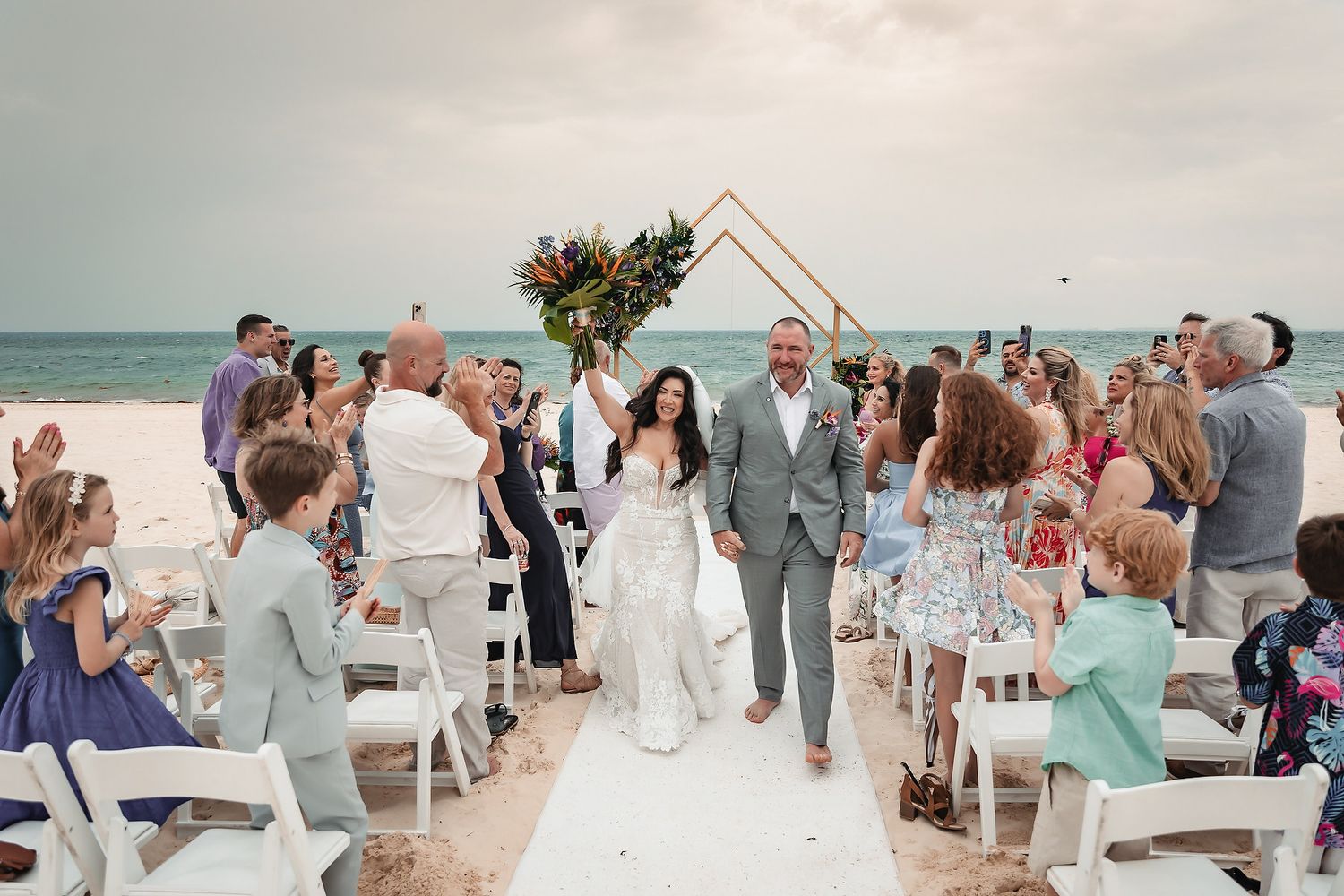Newlyweds walk down beach aisle surrounded by cheering guests under geometric floral arch at outdoor oceanside wedding ceremony.