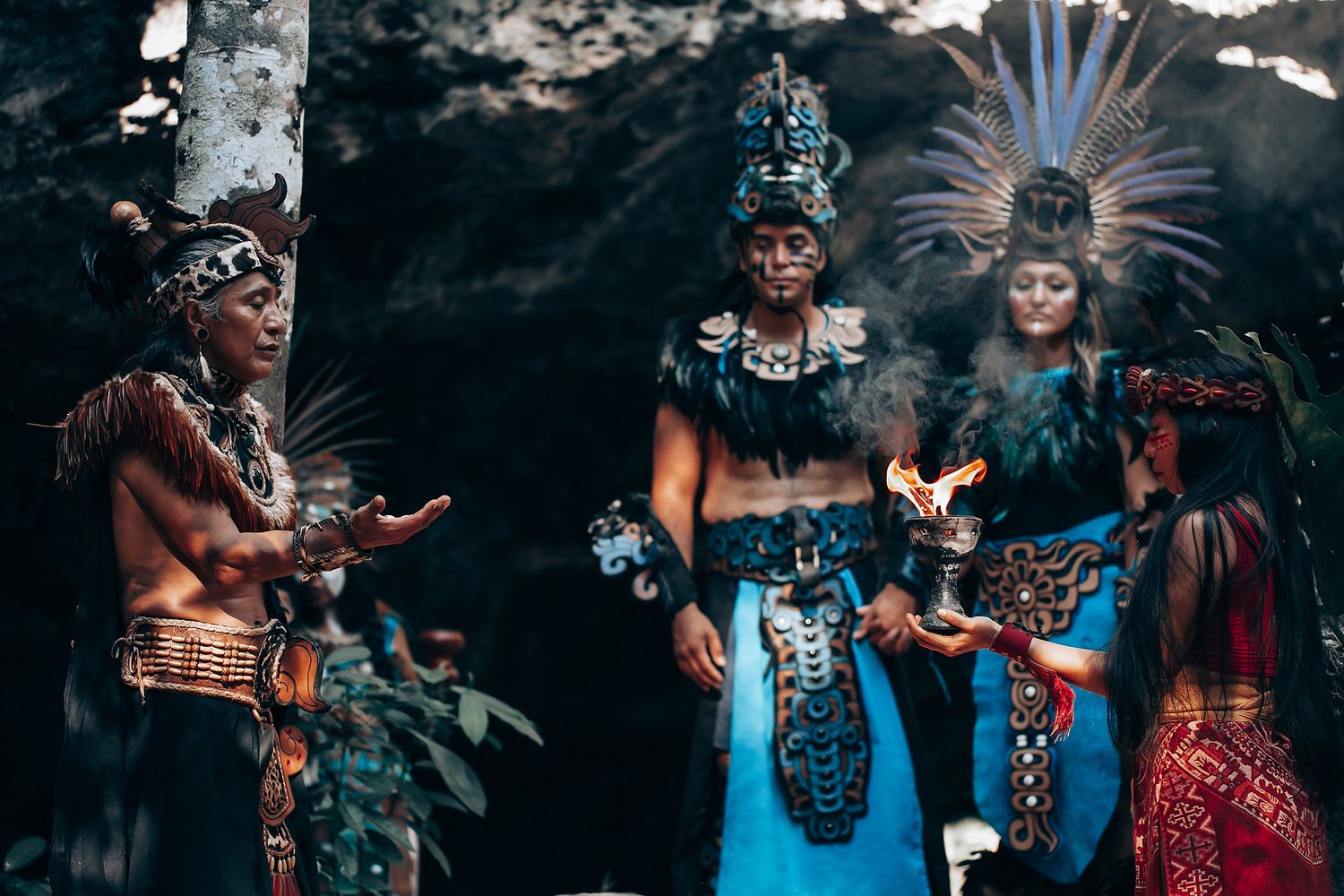 Group of performers in traditional Mayan ceremonial attire with feathered headdresses performing a fire ritual in a cave.