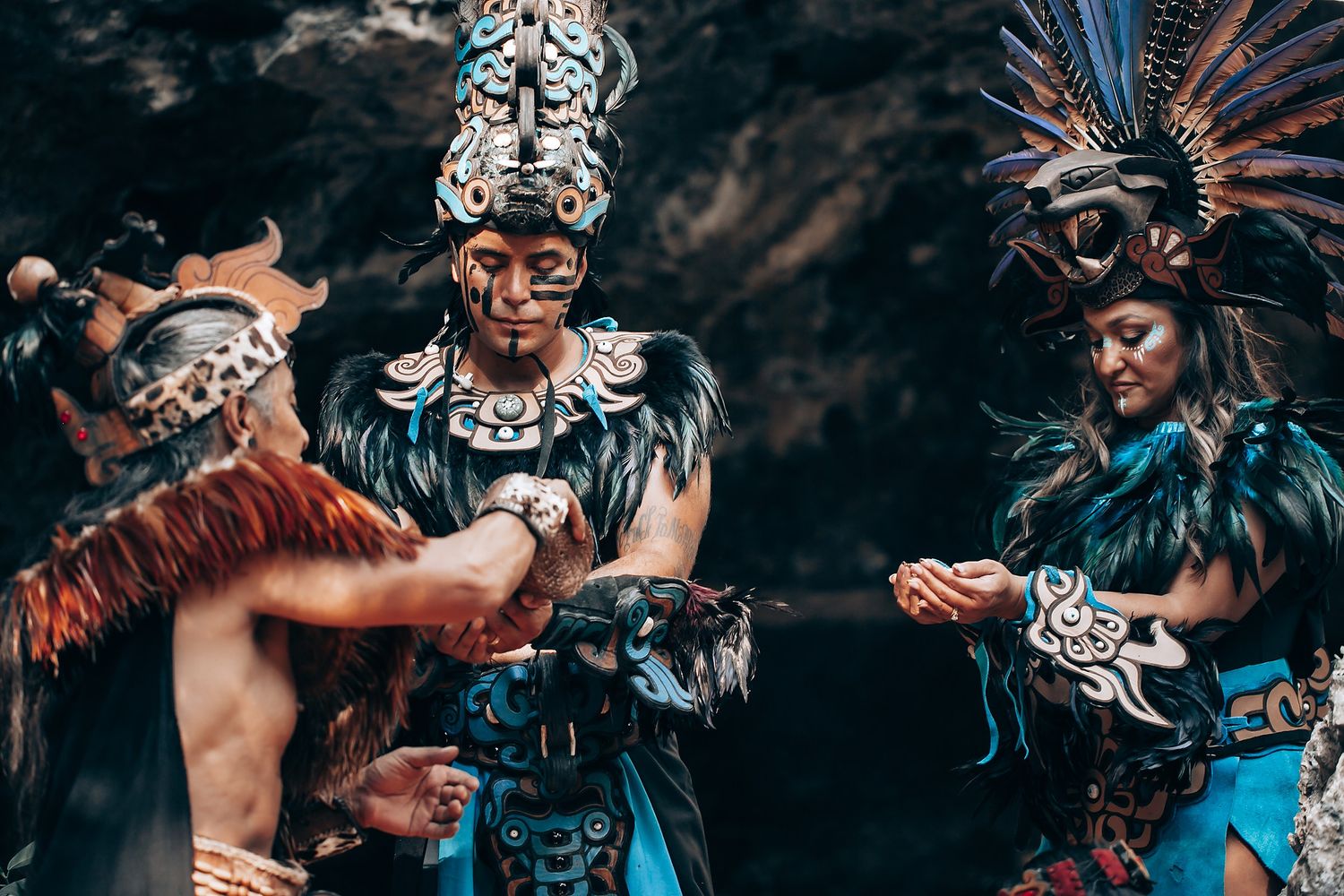Three Mayan ceremonial performers in elaborate feathered costumes and headdresses participate in a ritual near a cave.