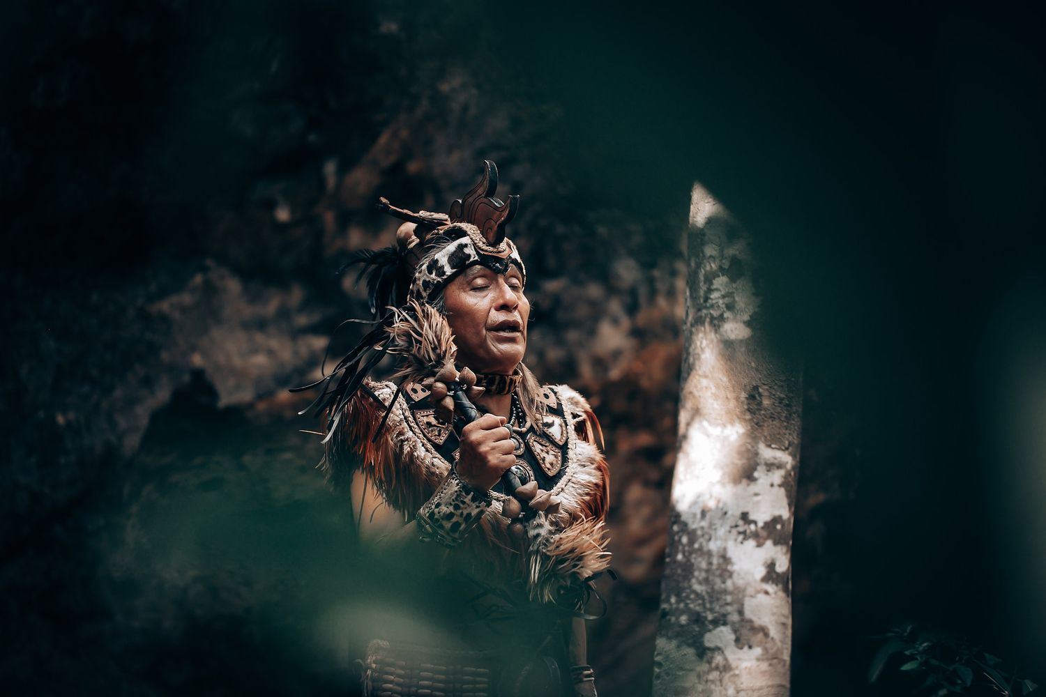 Indigenous person in traditional ceremonial attire with feathered headdress and beaded necklace standing in a lush forest.