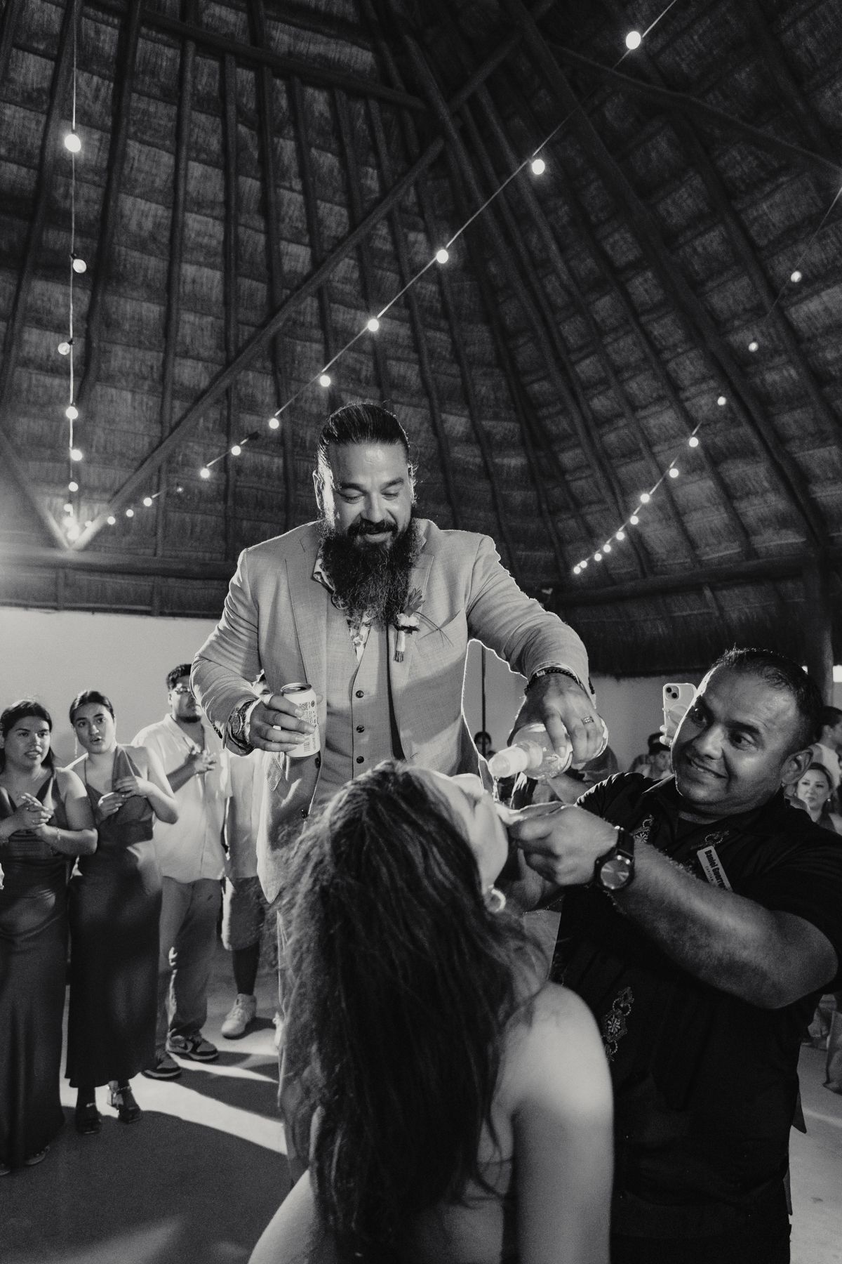 Groom lifted by guests during wedding reception celebration inside rustic venue with string lights overhead.