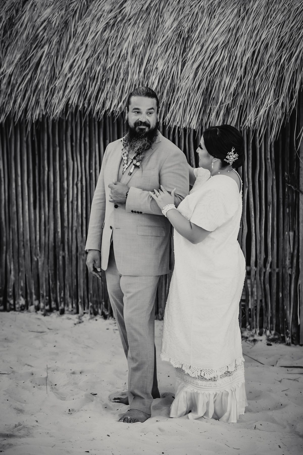 Black and white beach wedding photo of bearded groom in suit and bride in white dress standing on sand near thatched hut.