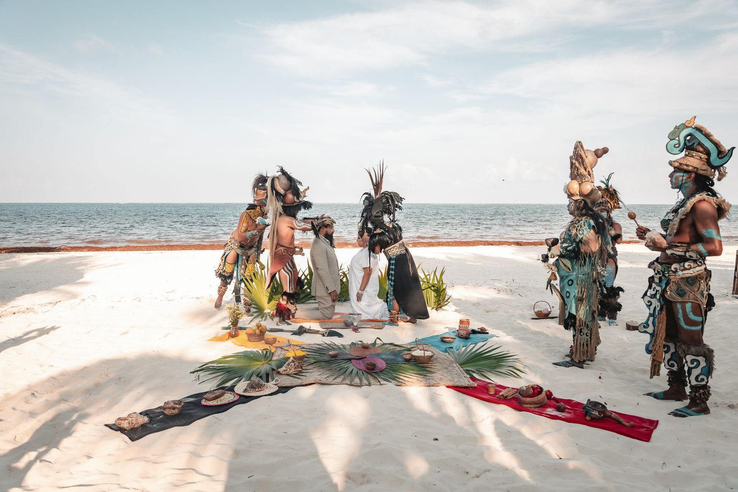 Mayan ceremonial performers in traditional costumes gather on a white sand beach for a ritual by the ocean.