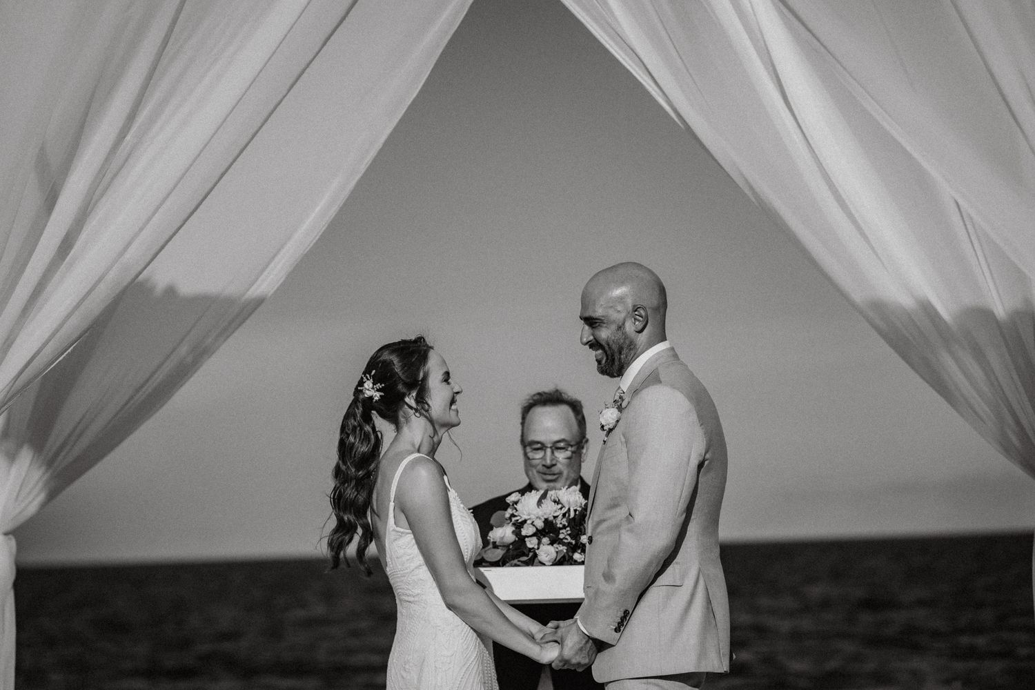 Black and white photo of bride and groom holding hands at beach wedding ceremony.