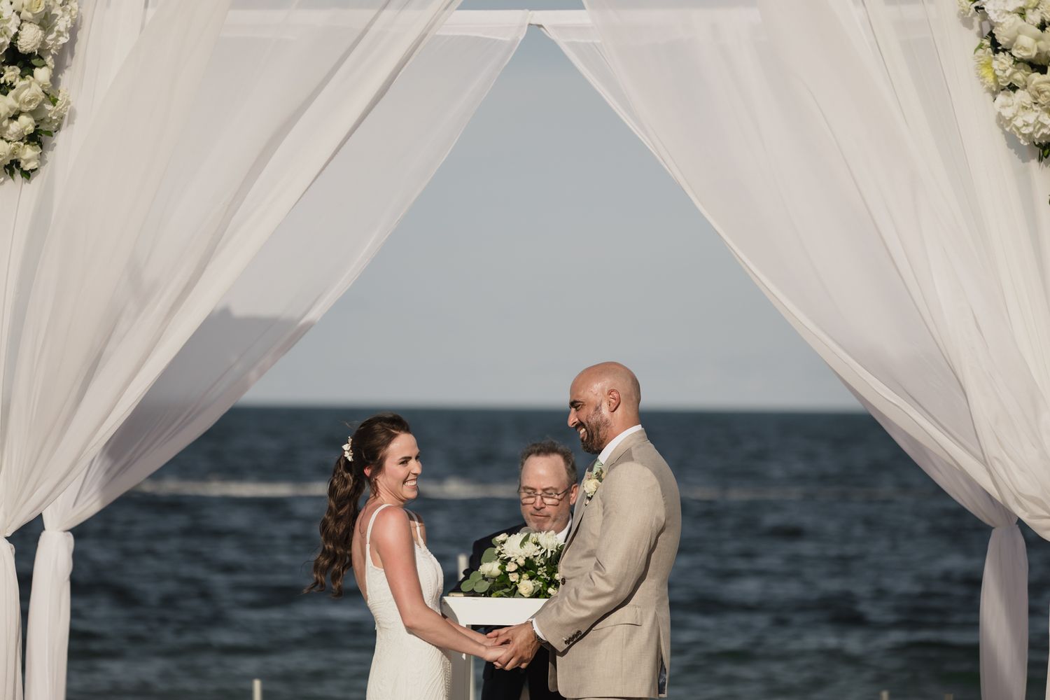 Couple exchanges vows at outdoor beach wedding ceremony under white draped arch with ocean backdrop.