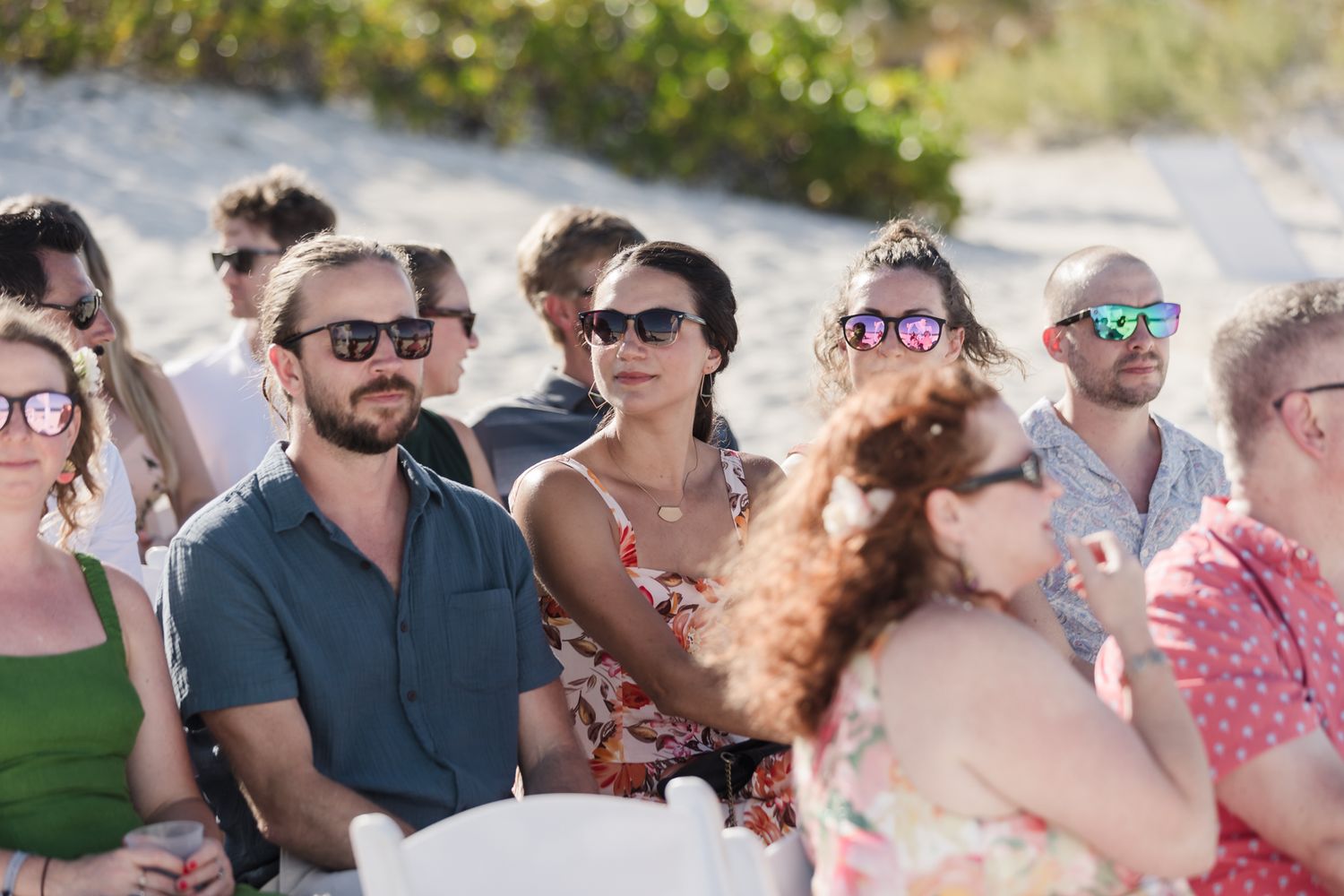 Guests in sunglasses seated outdoors at a beach wedding ceremony on a sunny day.