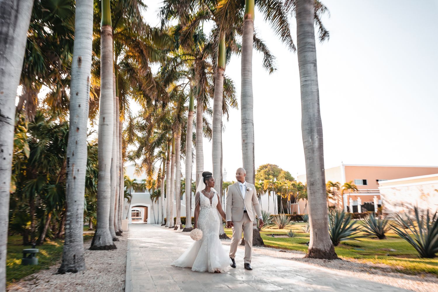 Bride and groom walk among tall royal palm trees at a tropical wedding venue during golden hour.