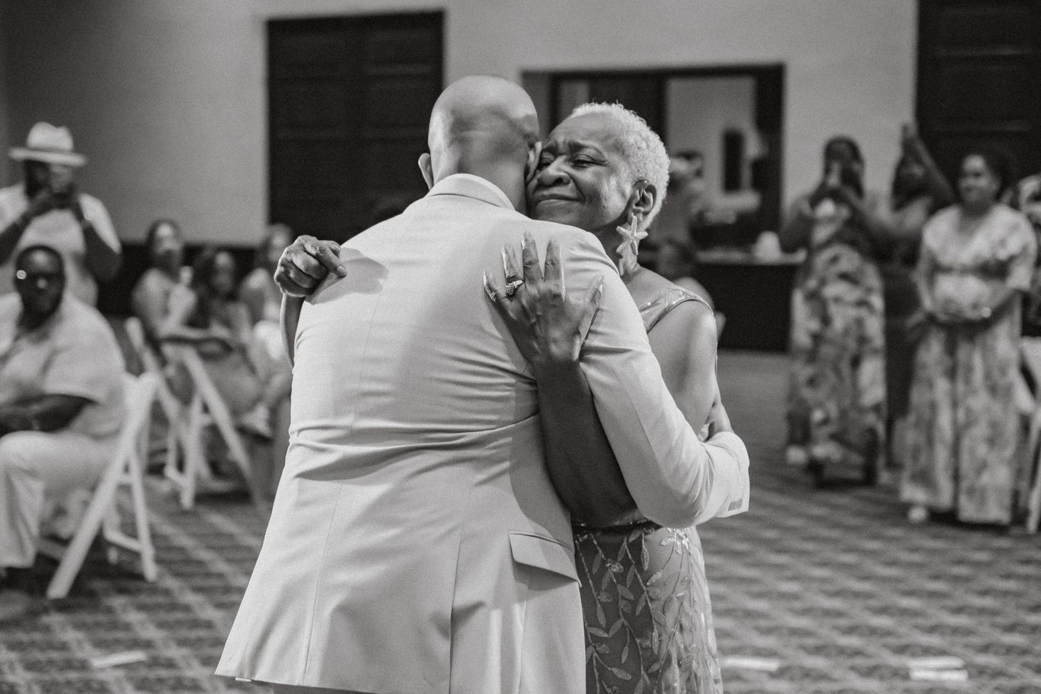 Groom shares tender dance with elderly woman at wedding reception in black and white photo.