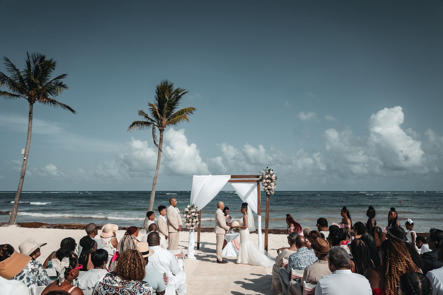 Beach wedding ceremony with couple at white arch, palm trees, ocean backdrop, and seated guests on sandy shore.