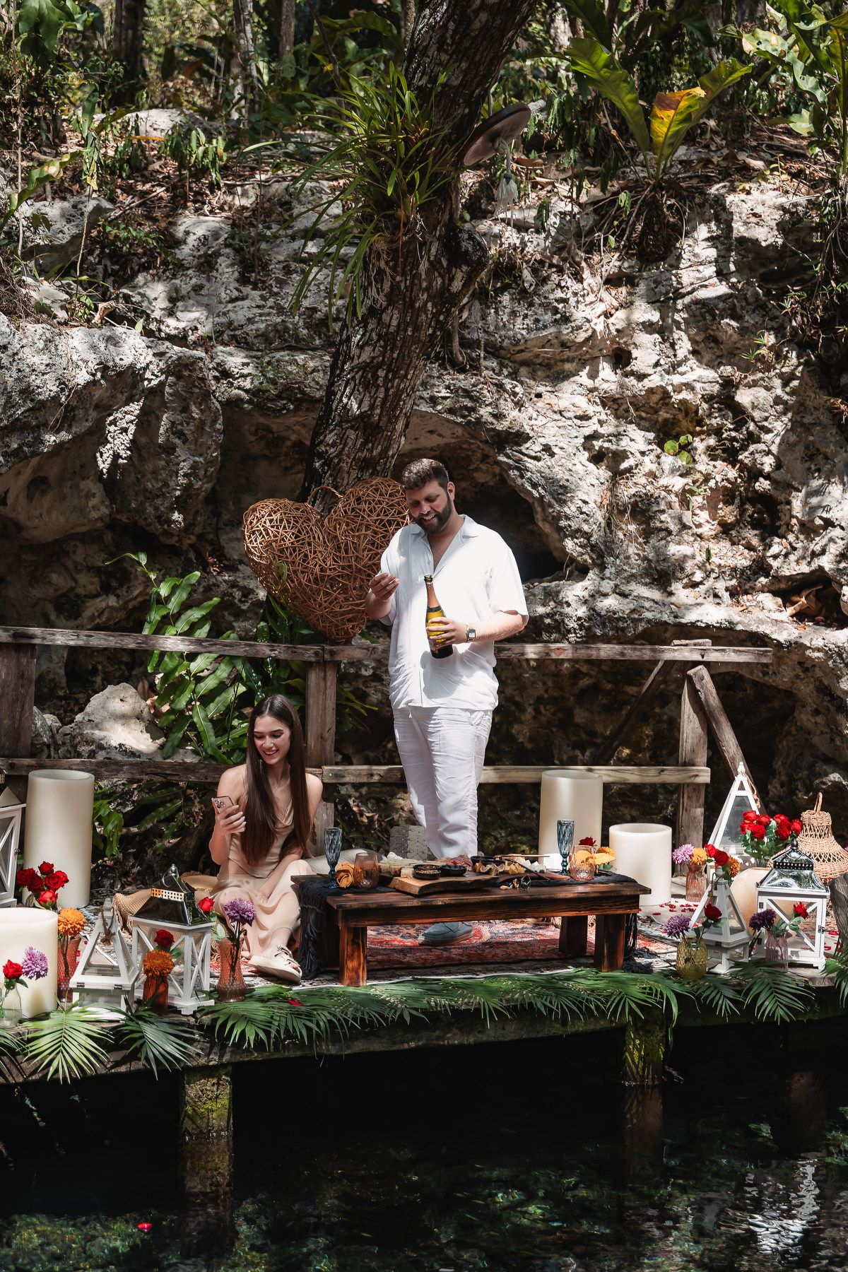 Man in white outfit holding champagne bottle beside woman at decorated table near a cenote in tropical jungle.