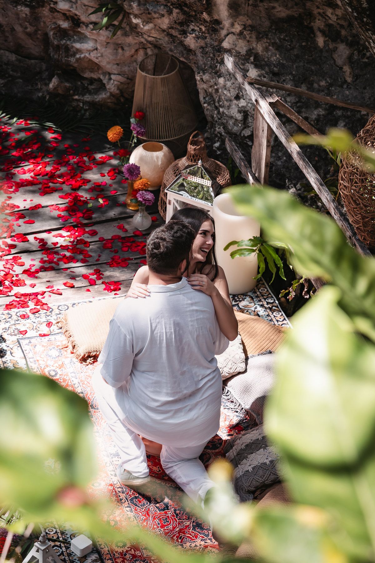Romantic couple embracing on a decorative rug surrounded by red rose petals and candles in a cave setting.