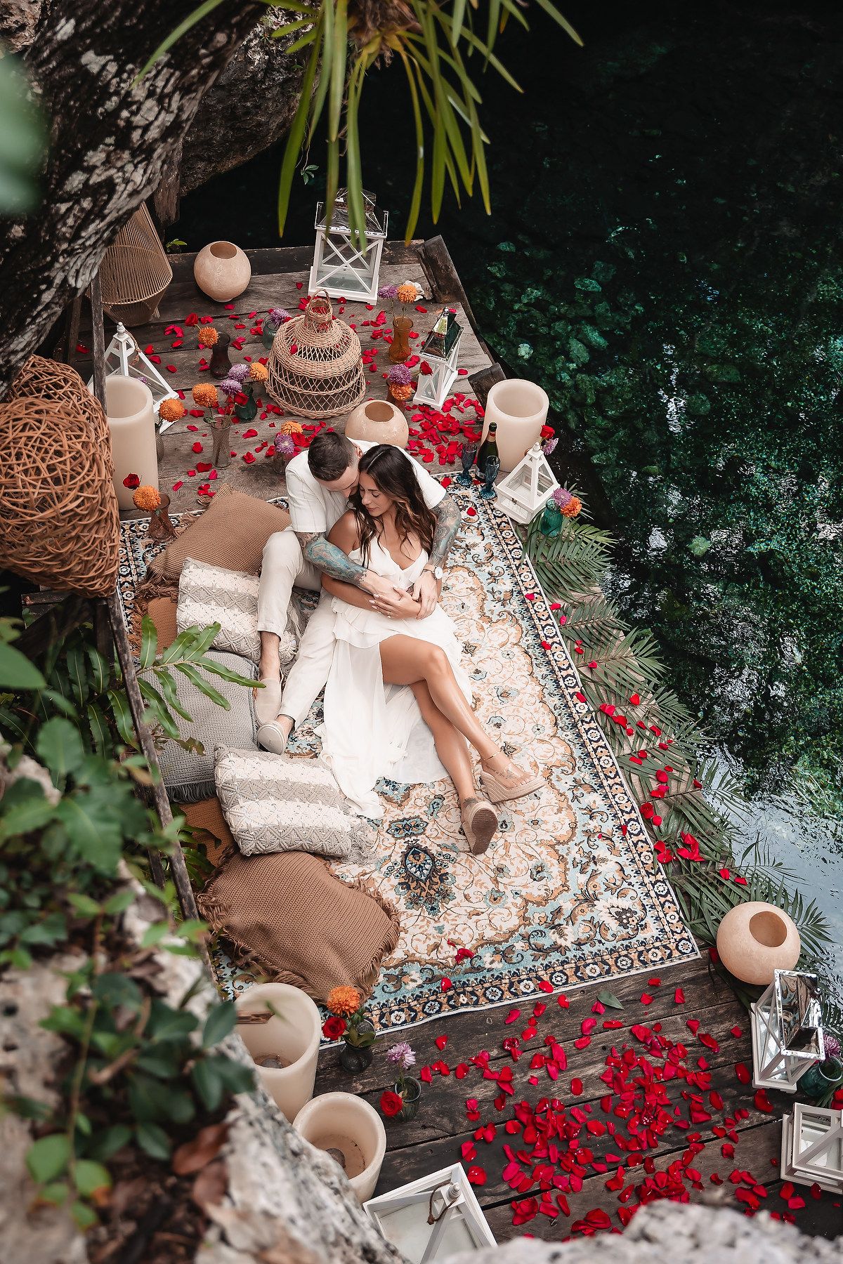 Couple relaxing on a boho-styled outdoor rug with rose petals, lanterns, and pillows by a serene jungle waterway.