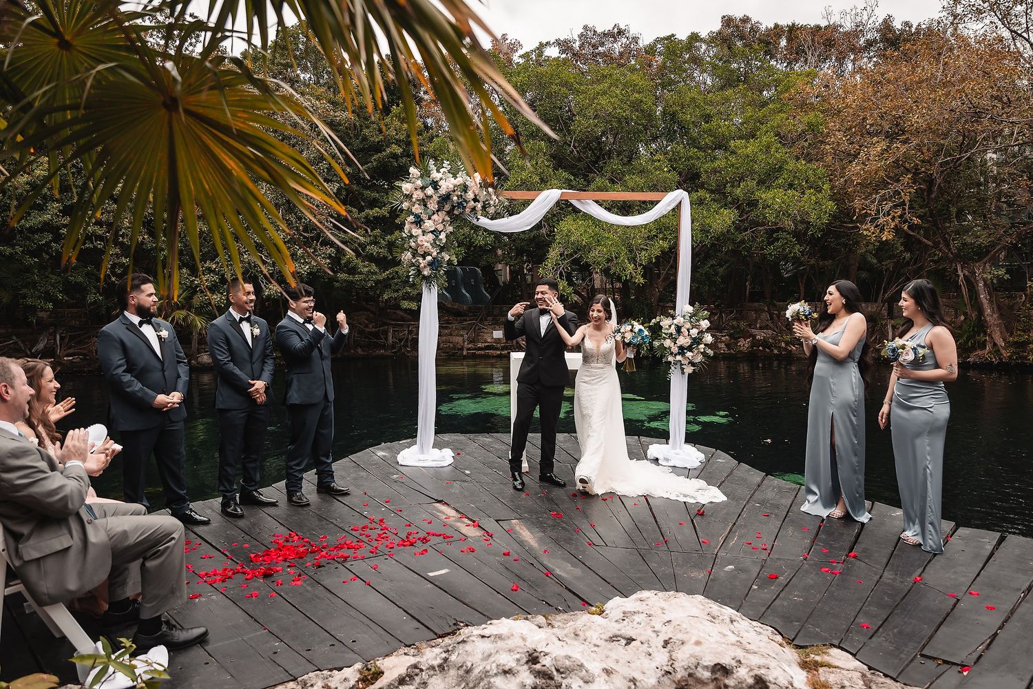 Outdoor wedding ceremony on a wooden dock by a lake, with floral arch, bridal party, and lush greenery backdrop.