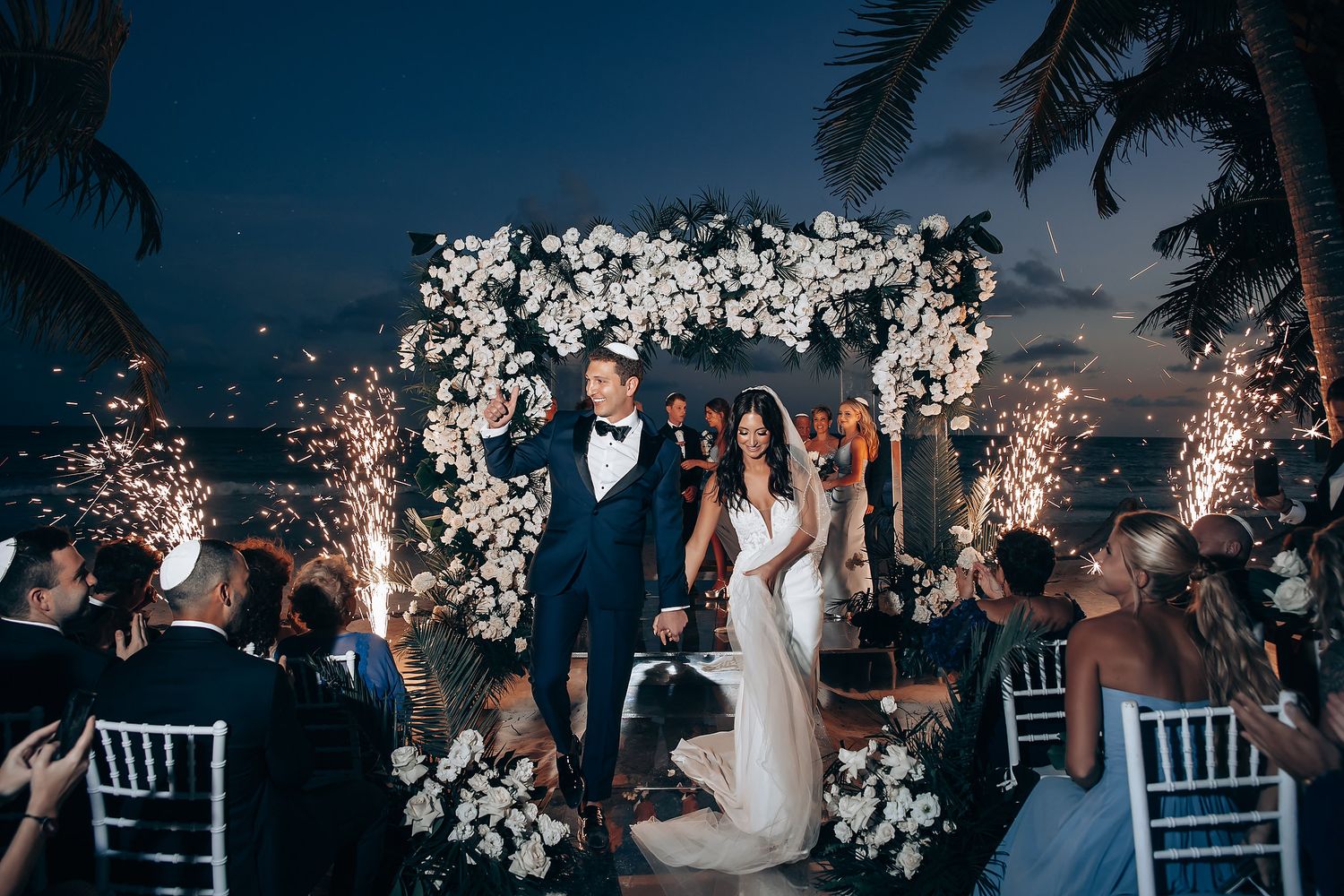 Evening beach wedding reception with sparklers and decorative lighting under palm trees.