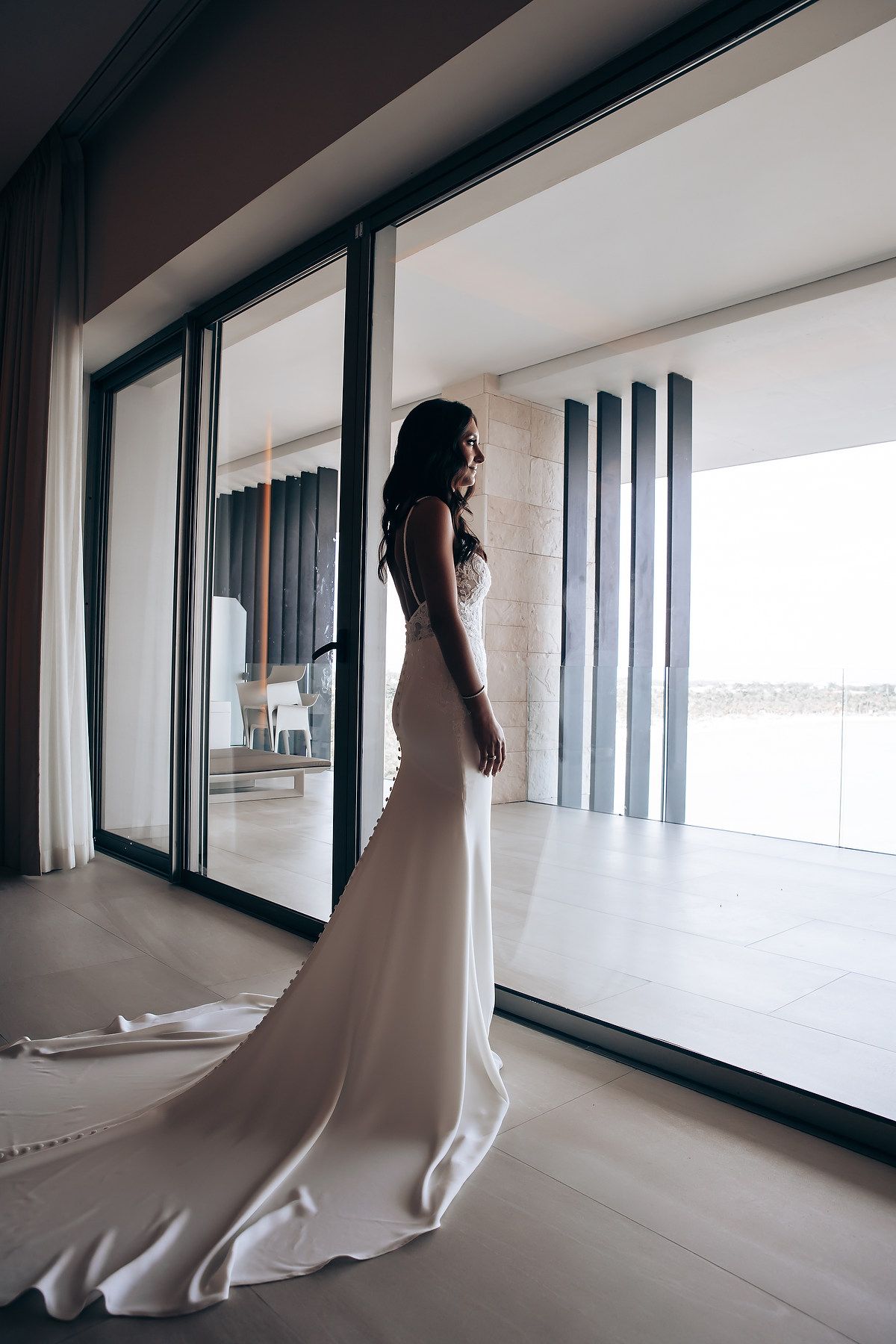 Bride in flowing white gown poses by large windows in modern architectural space.