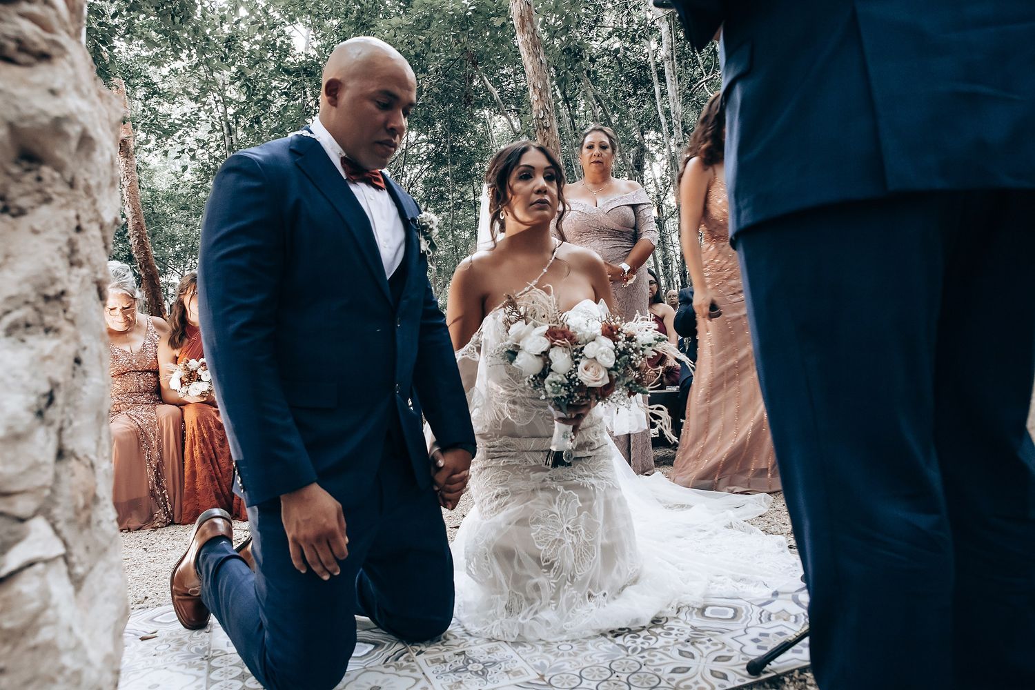 A couple kneels during an emotional outdoor wedding ceremony surrounded by guests and natural scenery.